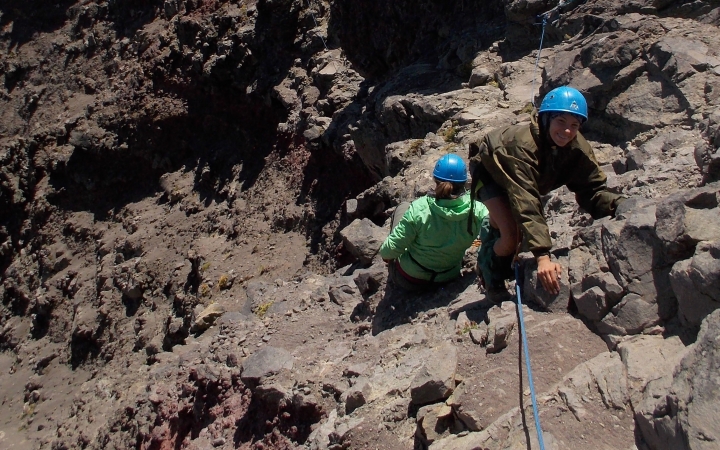 A person wearing rock climbing gear looks up at the camera and smiles while rock climbing. Below them, another person faces away from the camera.